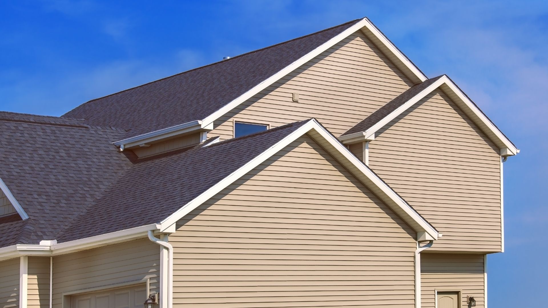 Modern suburban house with beige siding and dark gray roof against blue sky